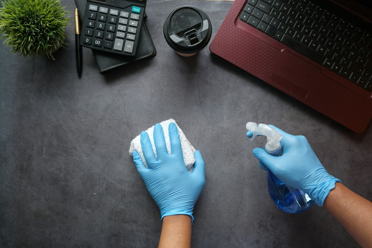 Handen in blauwe handschoenen maken een bureau schoon met een sproeifles en een doek
