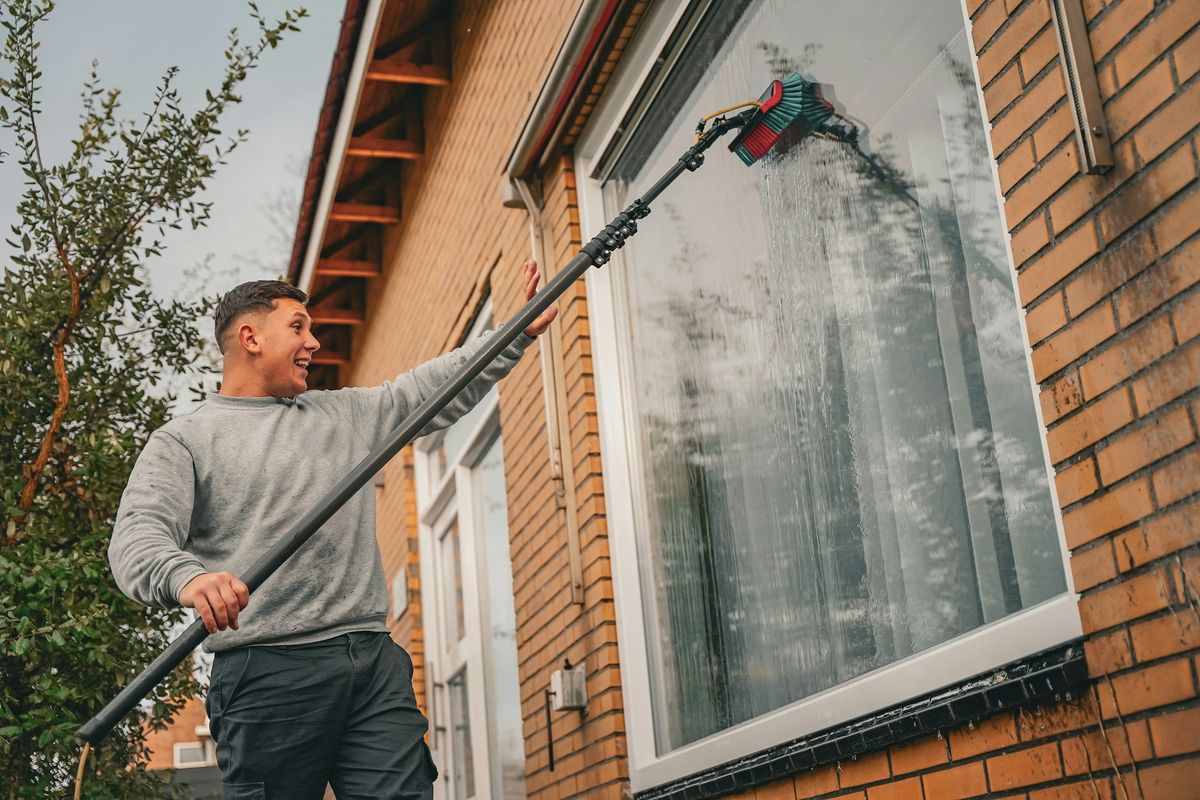 Een glazenwasser wast met een lange telescoopsteel het raam van een bakstenen huis